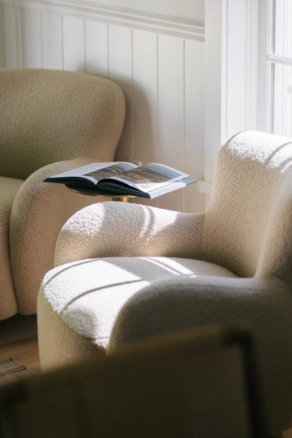 Close-up reading nook with boucle lounge chairs, small side table, and open book illuminated by soft afternoon sunlight